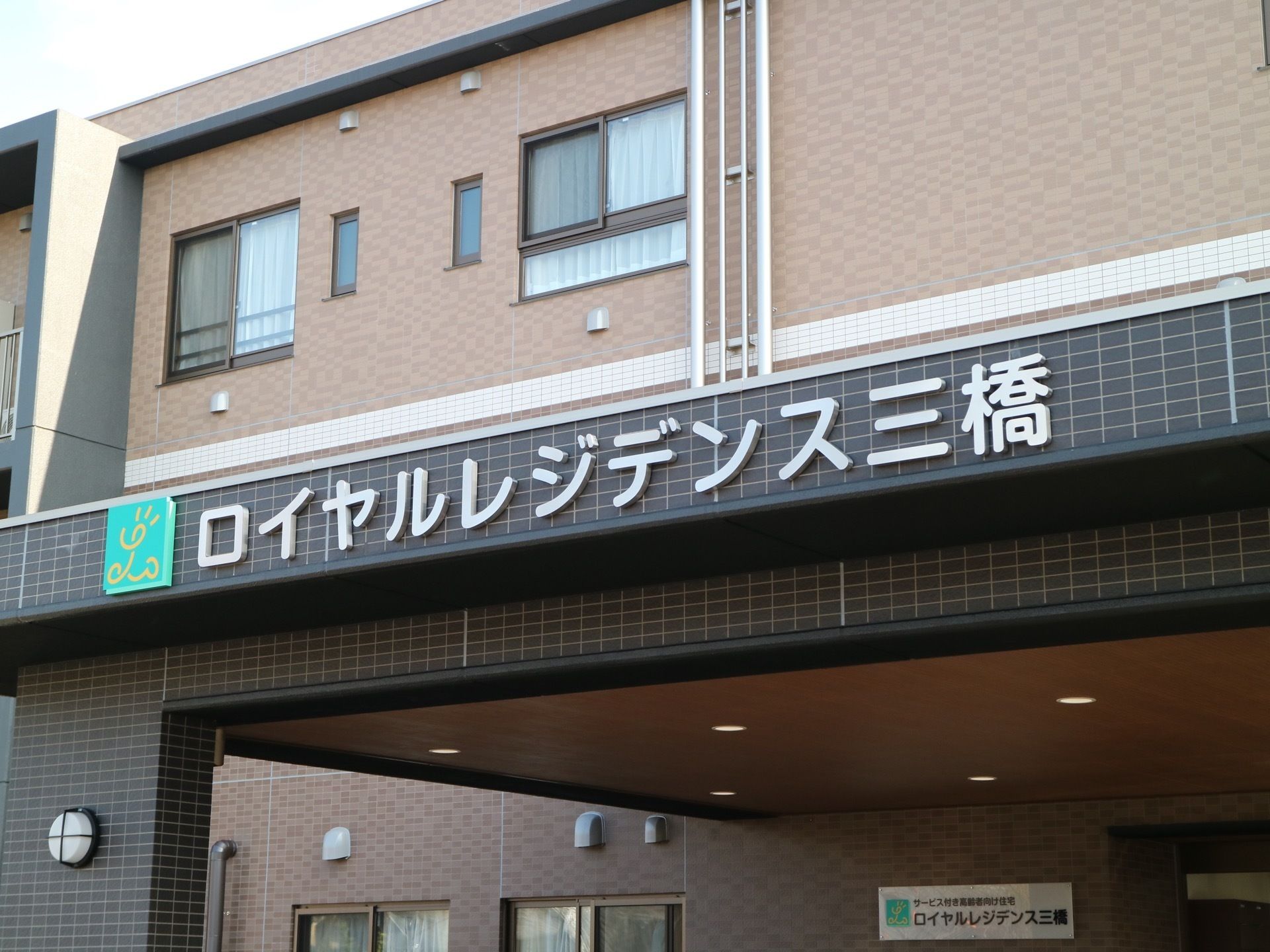 玄関から張り出しているひさしの部分に、施設名が掲示されている。その左端には緑色のロゴマークも入っている。