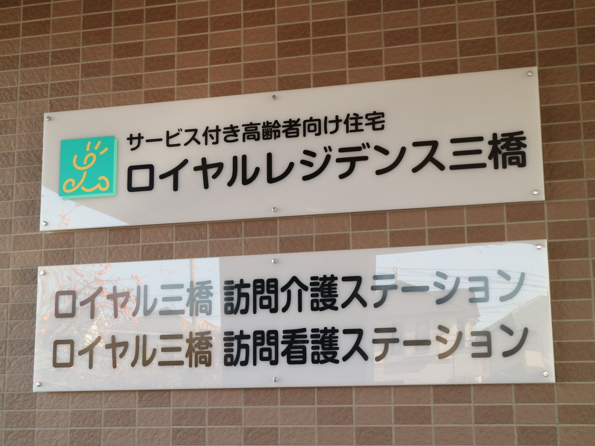 玄関の自動ドアの横には大きな看板が掲示されている。老人ホームのほか、訪問介護および看護サービス提供の旨が書かれている。
