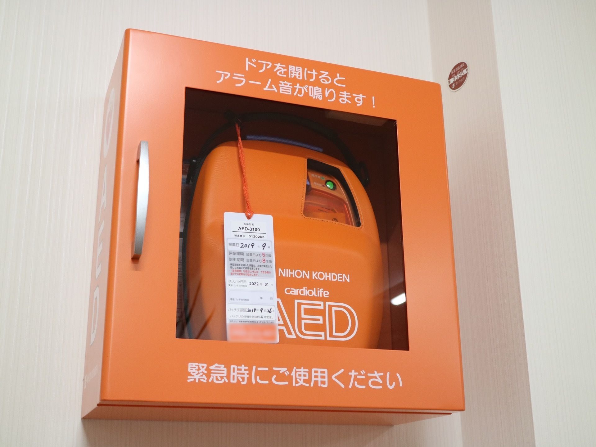オレンジのボックスに収納されているAEDで、本体も同じ色をしている。タグには設置年月が表示されている。