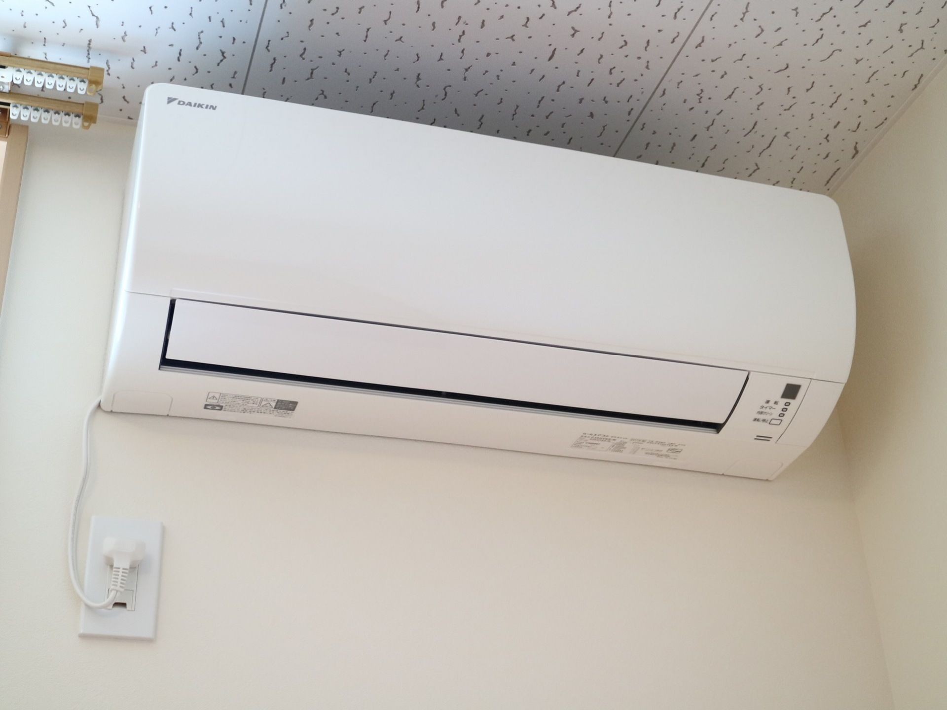 居室のカーテンレールの右側に、有名メーカーの白色のエアコンがある。電源コードが下に伸びて、コンセントに差し込まれている。