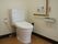 広いスペースに共用トイレがある。ふたつきの便座で、壁にはL字型の大きな手すりがかかっている。