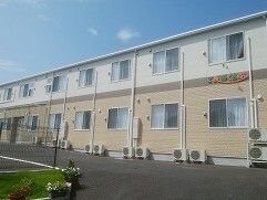 2階建ての建物は白とベージュでまとめられている。敷地内には駐車場があり、草木も植えられている。