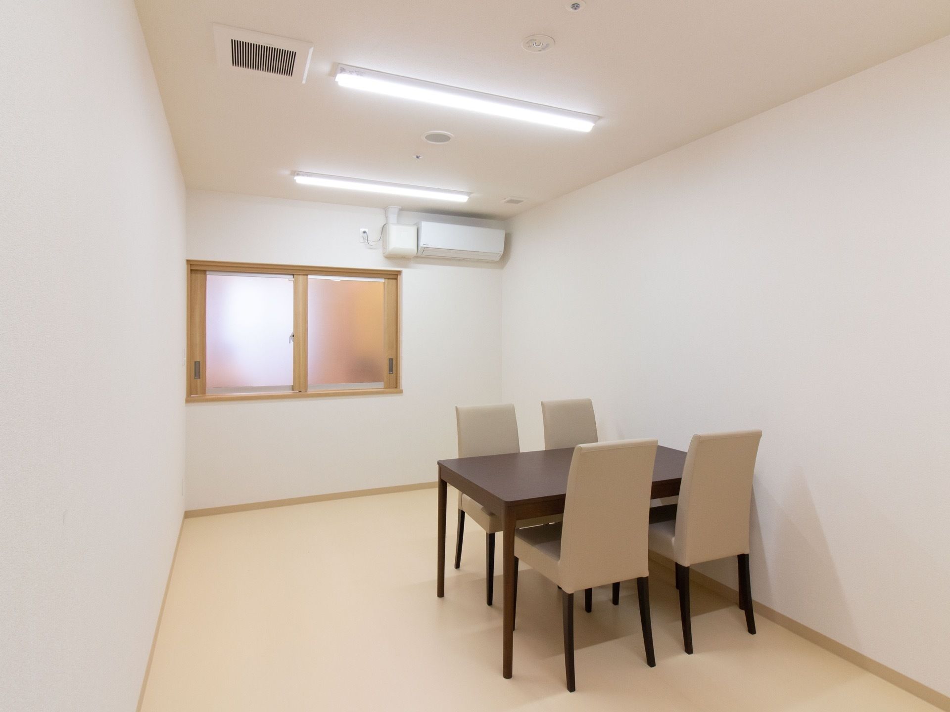 白を基調とした談話室の右側に、茶色いテーブルと白い椅子が設置されている。奥にはすりガラスの窓があり、壁にはエアコンが設備されている。