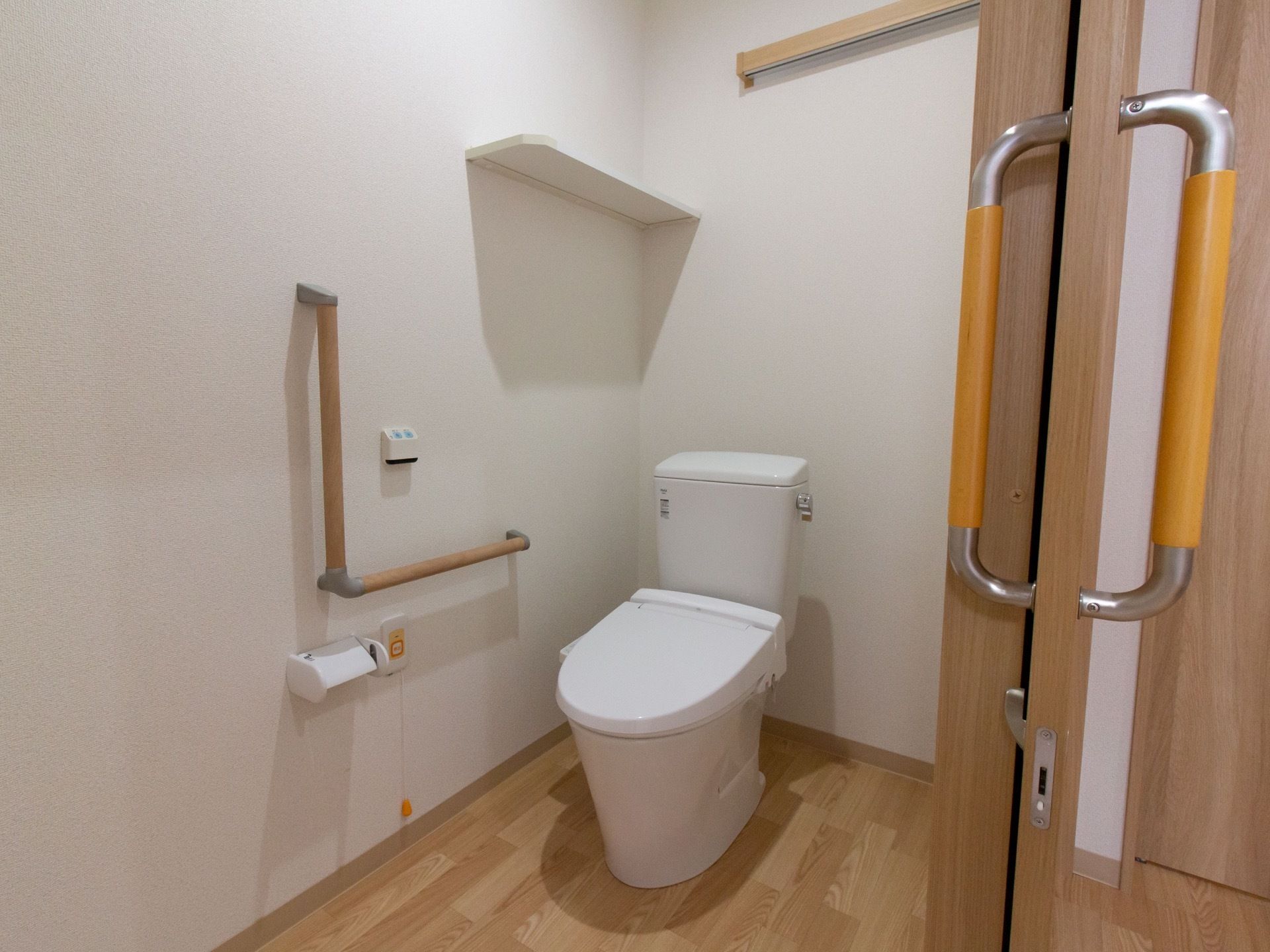 フローリングの床に水洗トイレが設置されている。壁には手すりやトイレットペーパーホルダー、呼び出しボタンがあり、上には棚がついている。