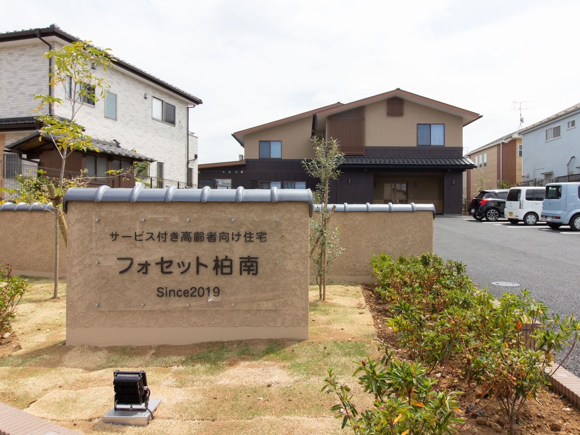 手前の花壇の中にある壁に施設名が書かれており、下にはライトが設置されている。奥には三角屋根の2階建ての建物があり、前には駐車場がある。