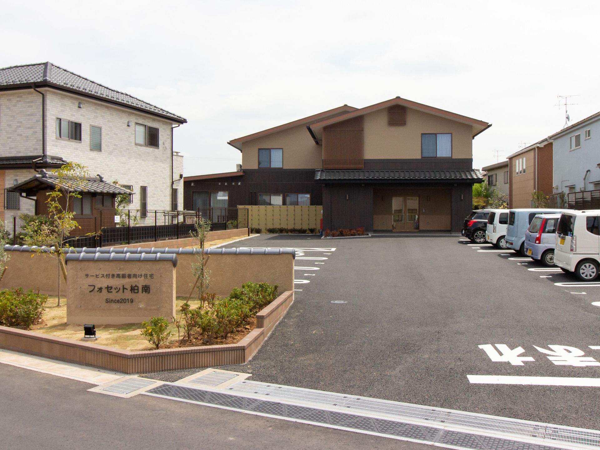 入口に茶色い看板と、緑の草が植わっている。奥の建物は茶色い壁と三角形の屋根が特徴だ