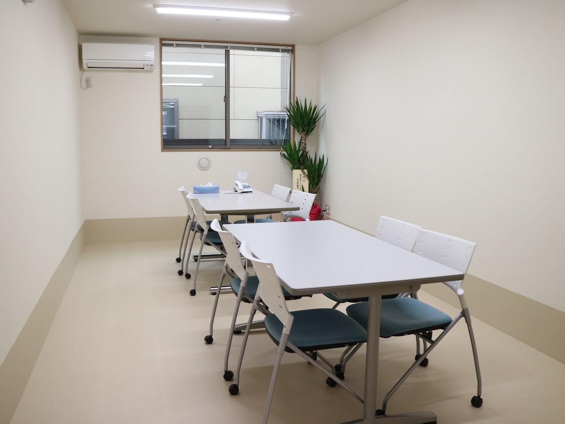 個室タイプの談話室で、奥に窓がある。窓の前には大きな観葉植物が置かれている。４人用のテーブルが２つある。