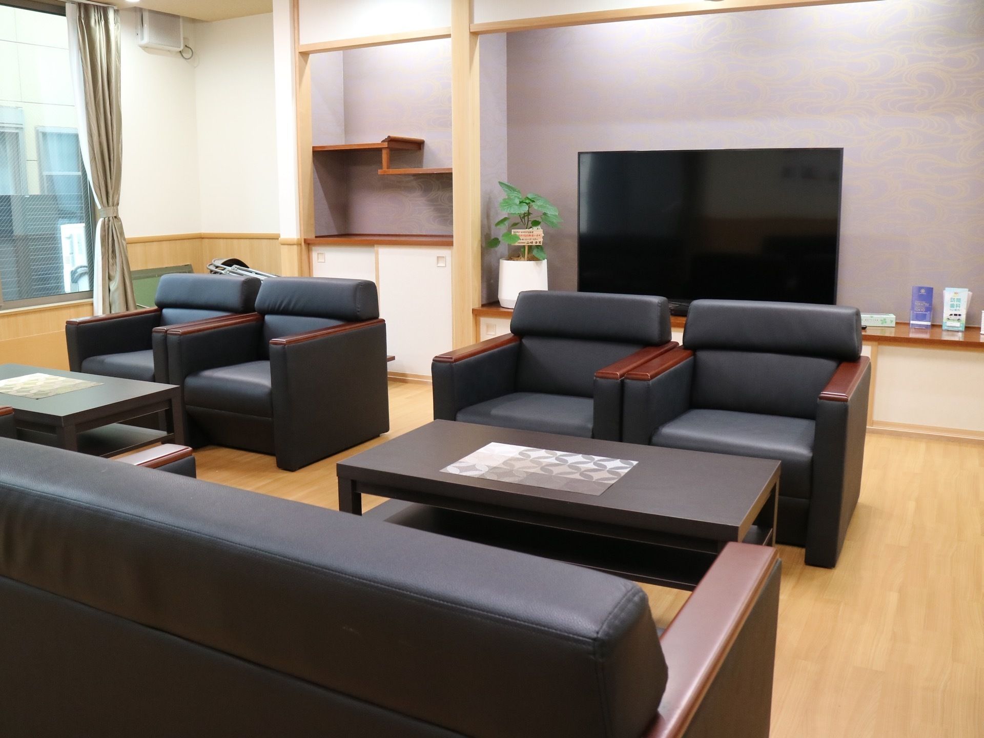 フローリングが広がる共有スペースで、床の間に大きな液晶テレビが置かれている。手前に黒いソファセットが並べられている。