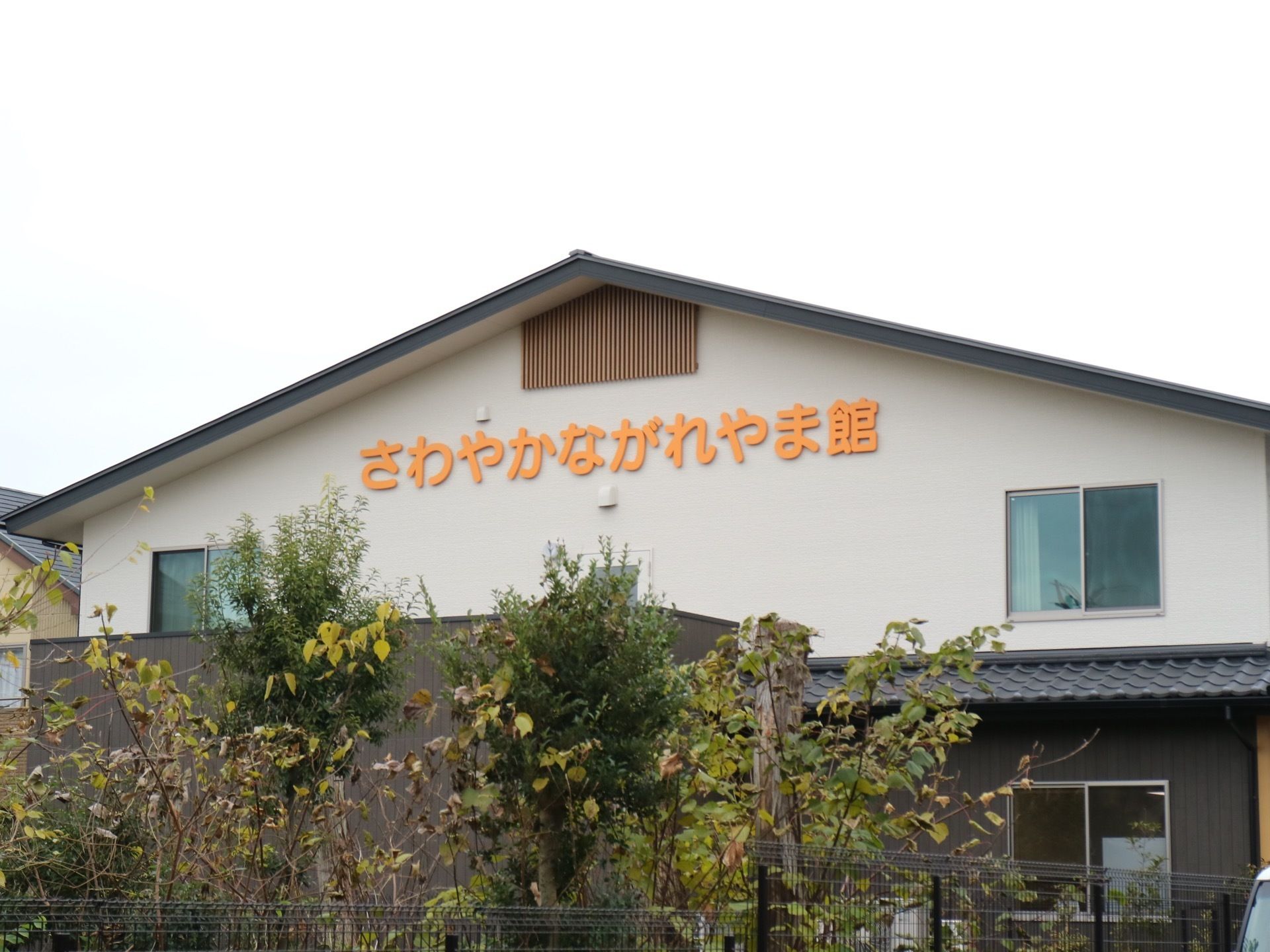 植栽に囲まれた敷地に、瓦屋根の建物が建っている。2階建てで、外壁には施設名のプレートが付けられている。