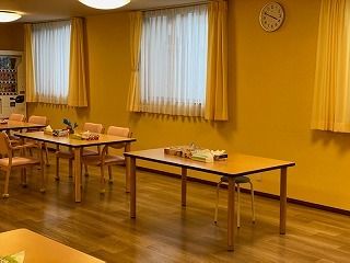 レースカーテンとカーテンが付いた大きな窓がある食堂には木製のダイニングテーブルが複数置かれている。