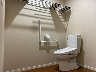 エル字型の手すりが設置されたトイレには棚やトイレットペーパーホルダーも付き、白でまとめられている。