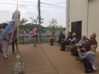 ウッドデッキのテラスに集まり、こいのぼりが泳ぐ姿の鑑賞している高齢者たち。スタッフが写真撮影をしている。