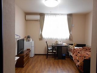 レースカーテンとカーテンが付いた大きな窓があるフローリングの部屋には寝具付きのベッドやテレビ、冷蔵庫などが置かれている。