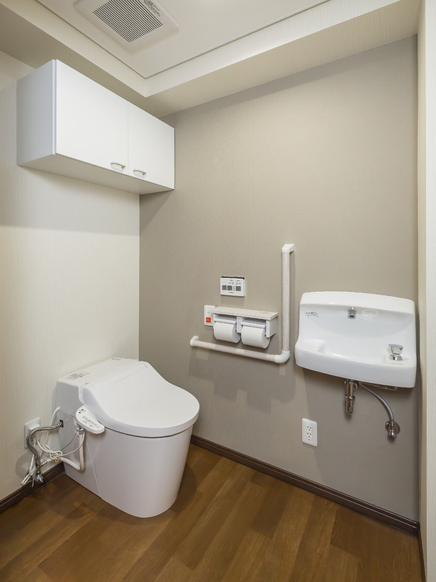 収納棚付きのトイレはフローリングでエル字型の手すりや手洗い場もある。