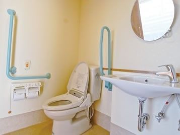 白でまとめられたトイレは広く、手洗い場付き。トイレの両サイドには水色の手すりが付いている。