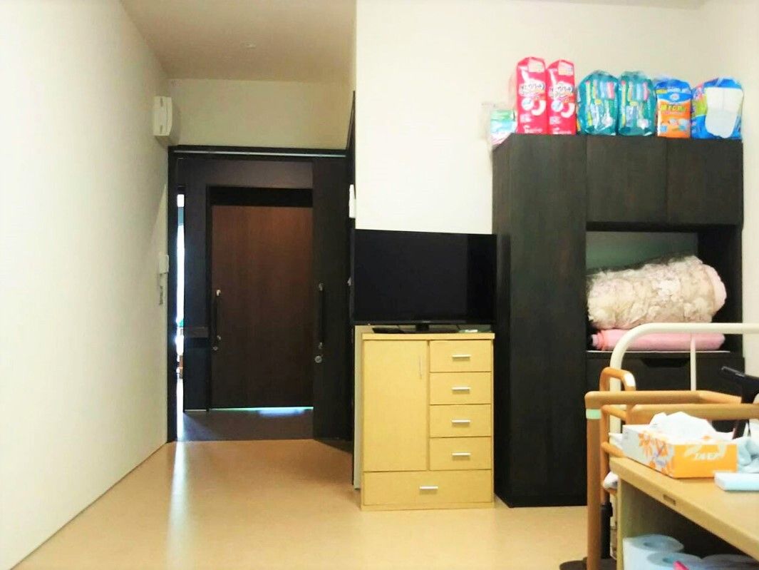 タンスやベッドなどが置かれた居室には大き目の黒い棚もあり、その上にはおむつなどが並べられている。出入り口はスライド式のドア。
