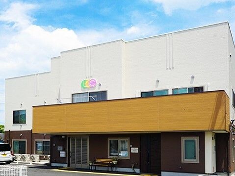 白とブラウン系の落ち着いた雰囲気の建物は2階建て。敷地内には駐車スペースがあり、玄関先にはベンチも置かれている。