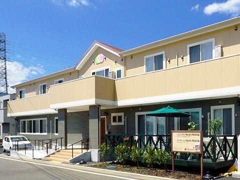 駐車場やベランダ、テラスが付いたベージュの２階建ての建物。窓がたくさんつき、１階部分の出入り口には屋根と階段、スロープが付いている。