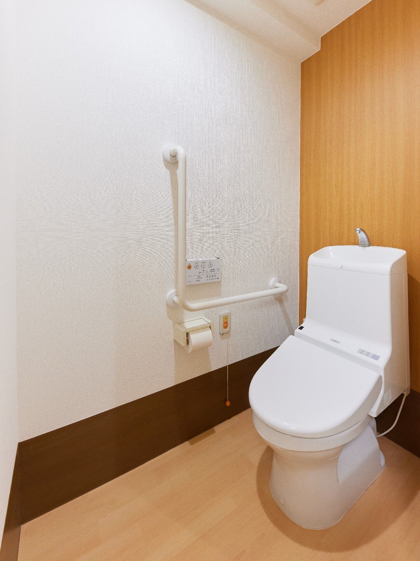 エル字型の手すりやオレンジの呼び出しボタンが付いたトイレはい広く、温水便座が付いている。