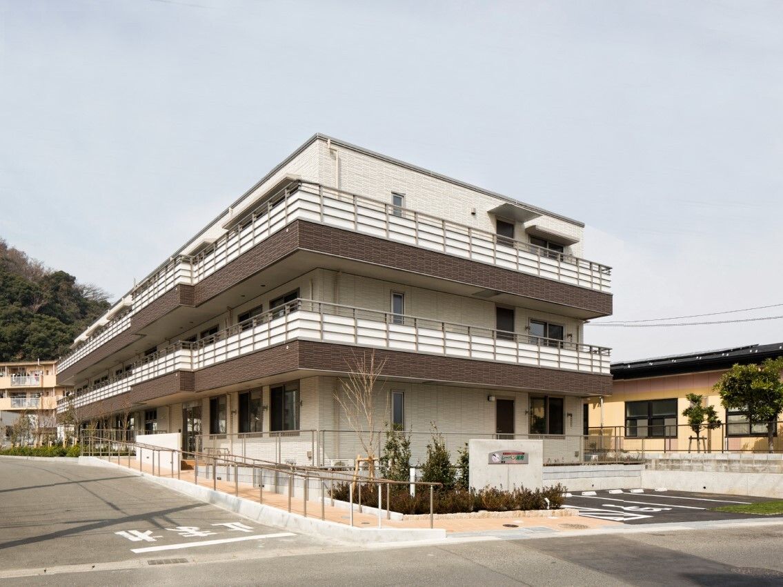 ベランダ付きの3階建ての建物。エントランスにはスロープが付き、花や木が植えられている。