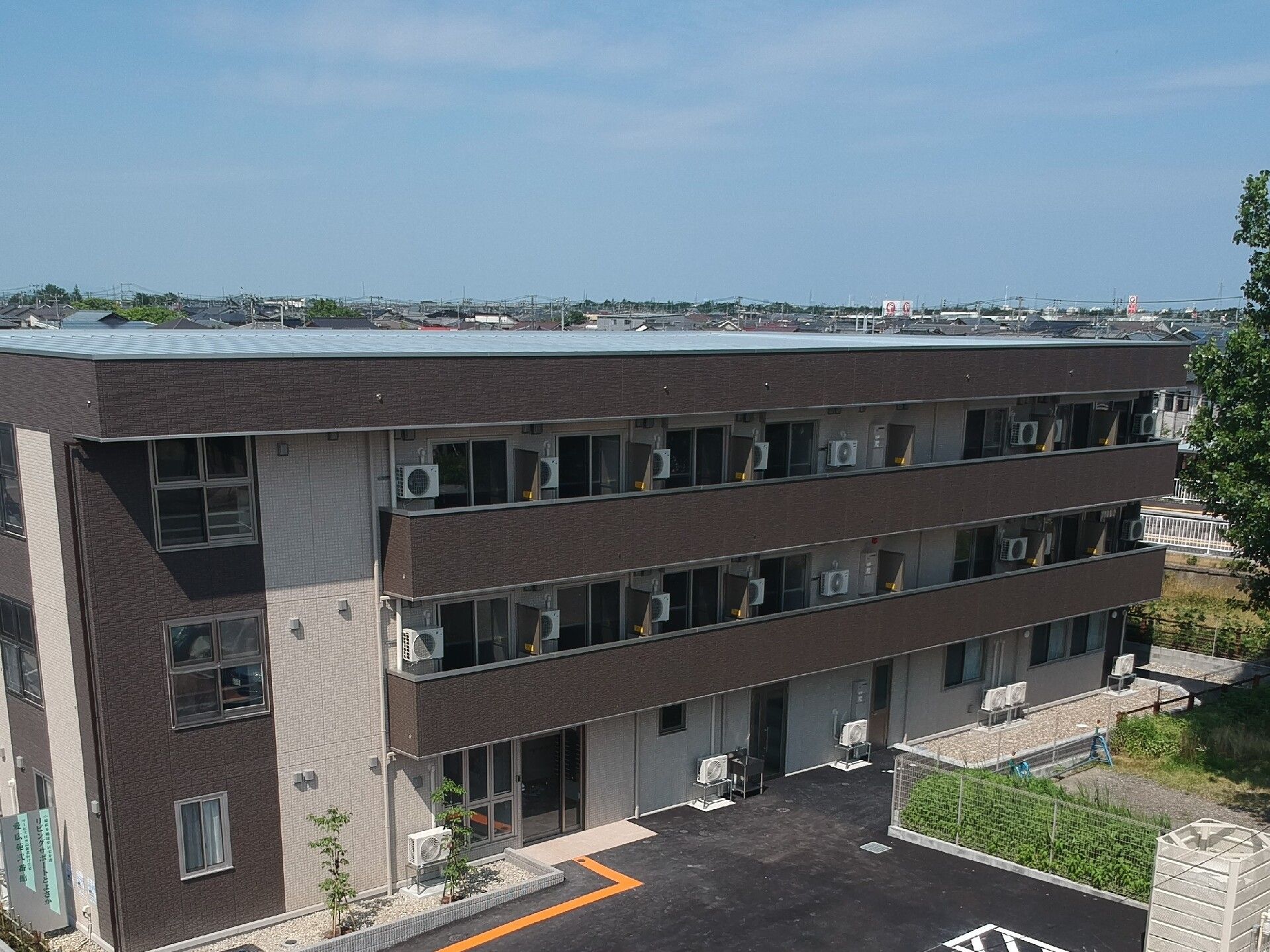 ブラウン系の3階建ての建物。広い駐車場があり、点字ブロックが敷かれている。緑が植えられた場所もある。
