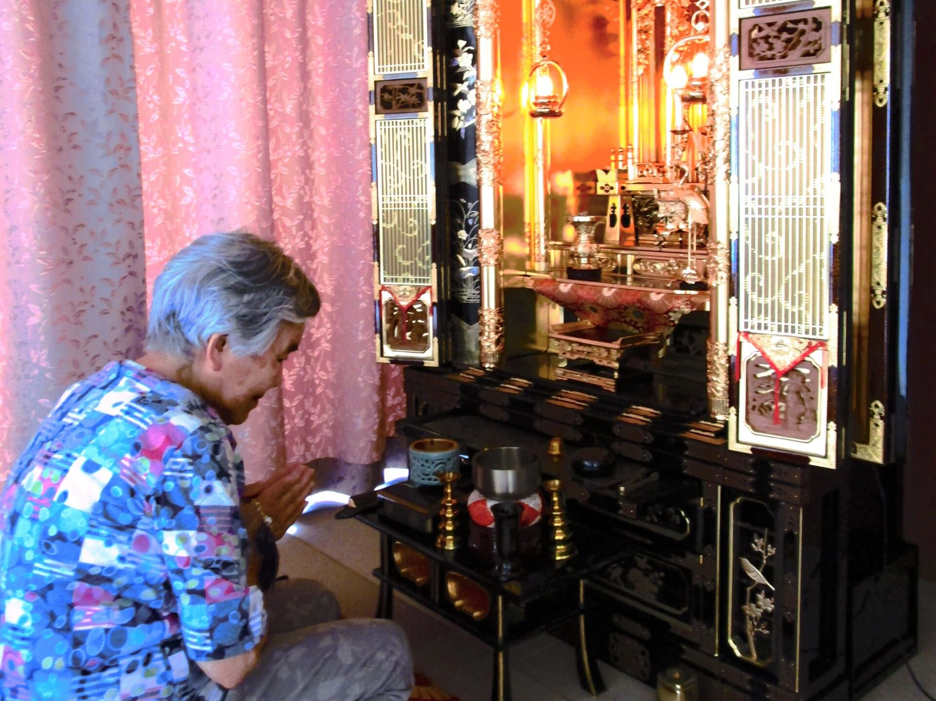 ピンク色のカーテンが閉められている部屋で仏壇に向かって手を合わせている人
