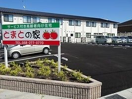 建物名が書かれた館銘板が設置された小さな花壇スペースが手前にあり、その後ろには広い駐車場や住宅の建物がある。