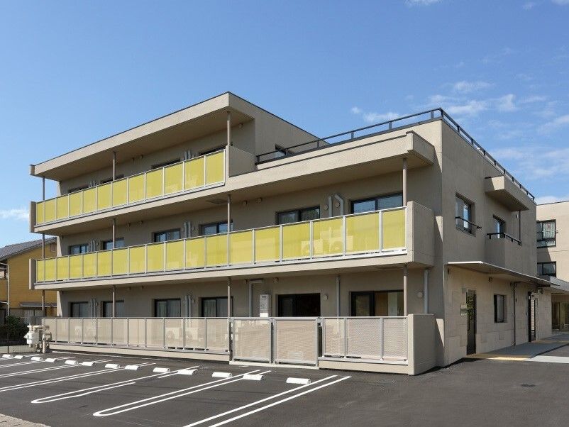 広い駐車場が付いた3階建ての建物の2階部分と3階部分にはベランダがあり、黄色い柵が付いている。
