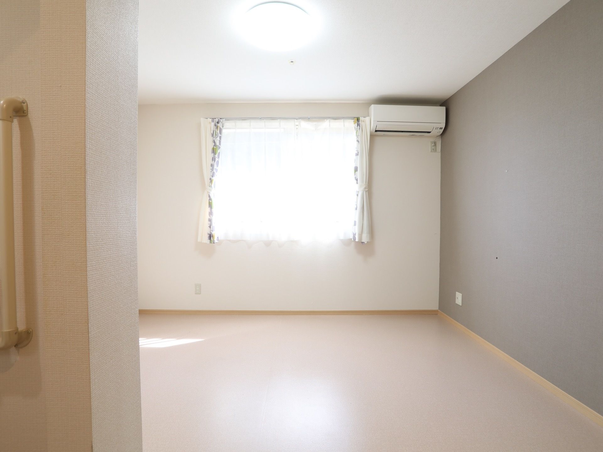 エアコンが備わっている居室である。窓にはレースのカーテンとドレープカーテンが下がっている。明るい日差しが差し込んでいる。