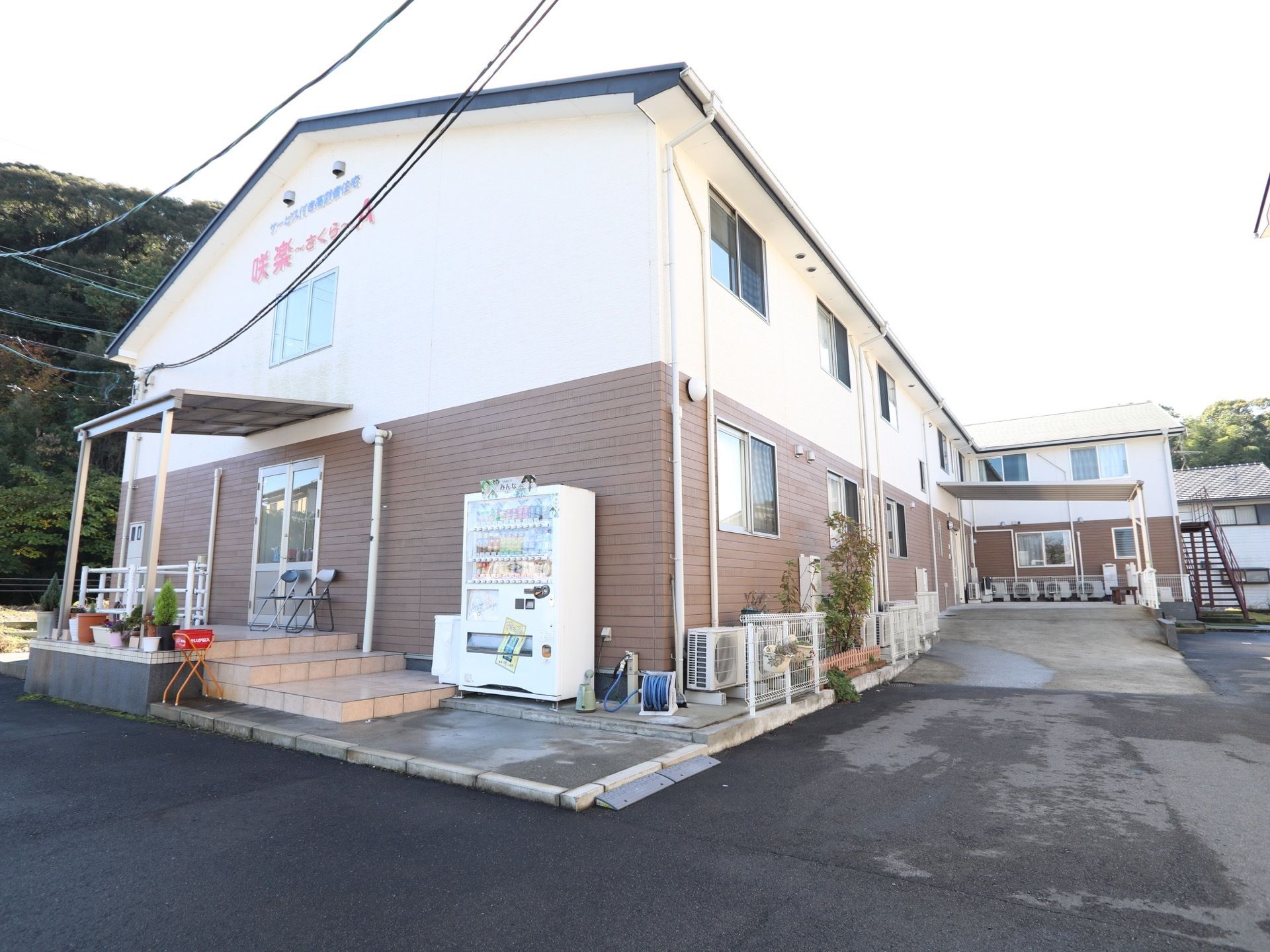 高い建物は周辺になく、近い場所に鬱蒼と茂った木々がみられる。ひさしの下に、パイプ椅子が２個並べて置かれている。