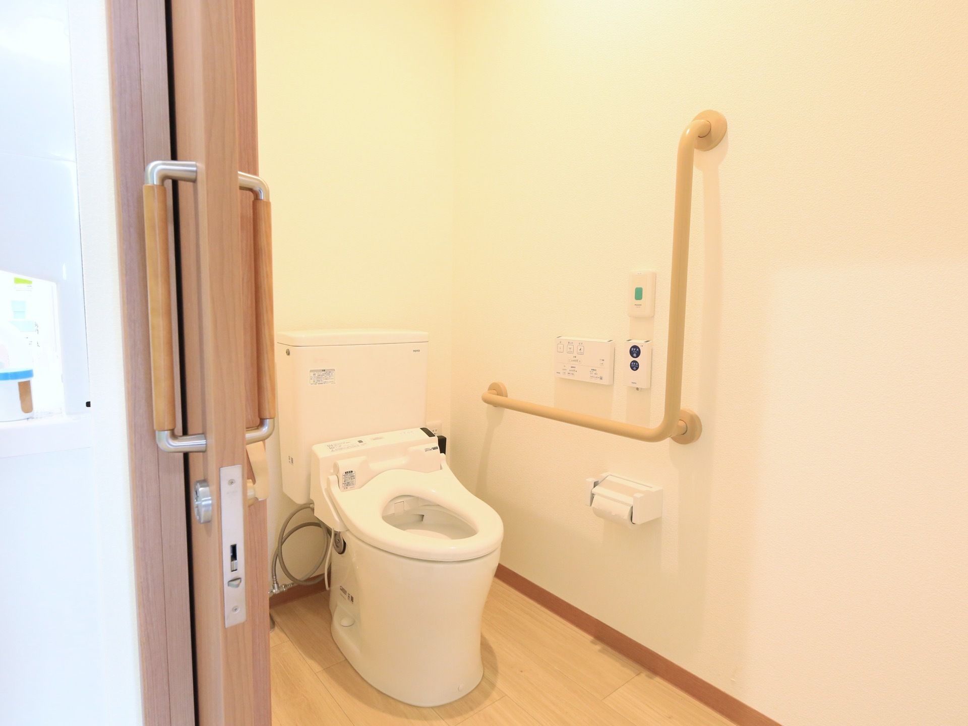 扉左手奥に、温水洗浄便座のトイレがある。壁にＬ字の手すりとペーパーホルダー、操作パネル、緊急ボタンがある。