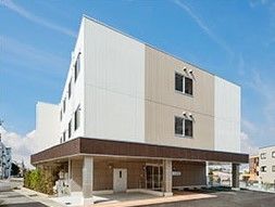 白とベージュの3階建ての建物。1階部分には数台分の駐車スペースもあり、木も植えられている。