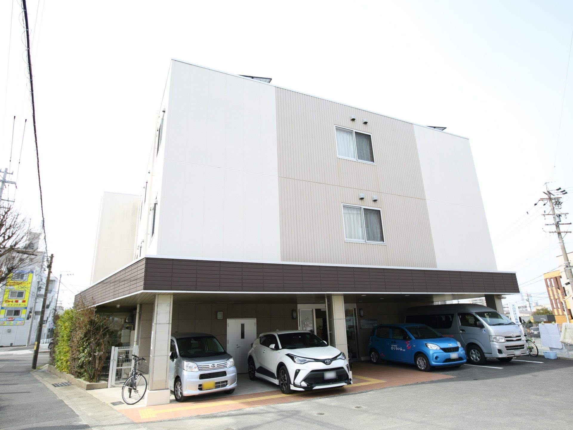 白とグレーで塗り分けられた建物である。中央に窓があり、その下の１階部分は駐車スペースになっている。車が４台停められている。