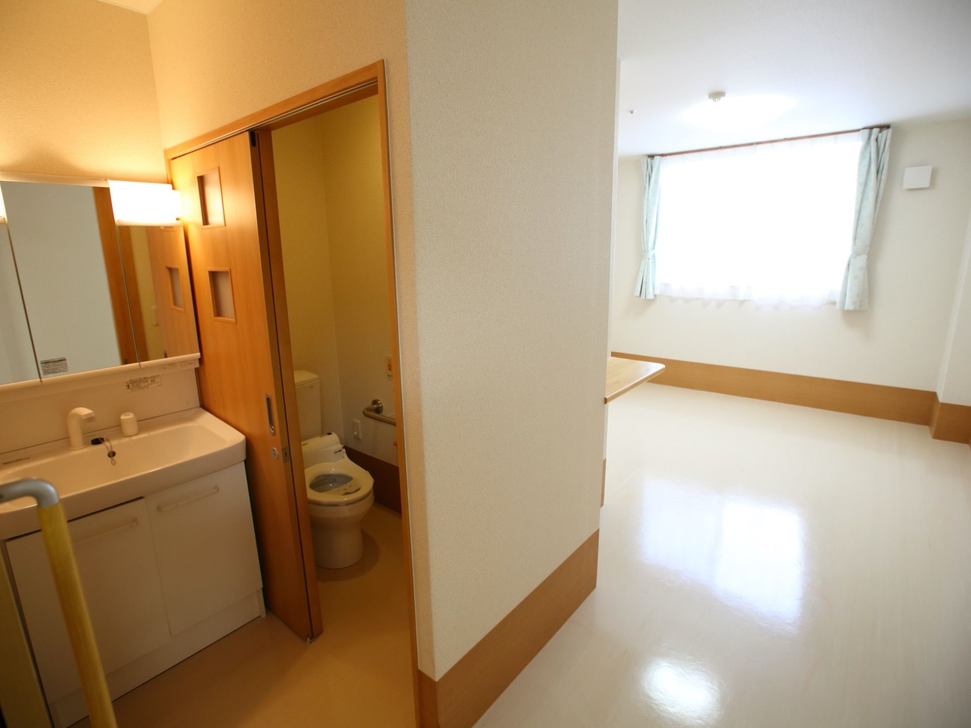 居室内のトイレと洗面台の画像である。右奥に居住スペースが広がり、左手前にトイレと洗面台がある。居住スペースに大きな窓がある。