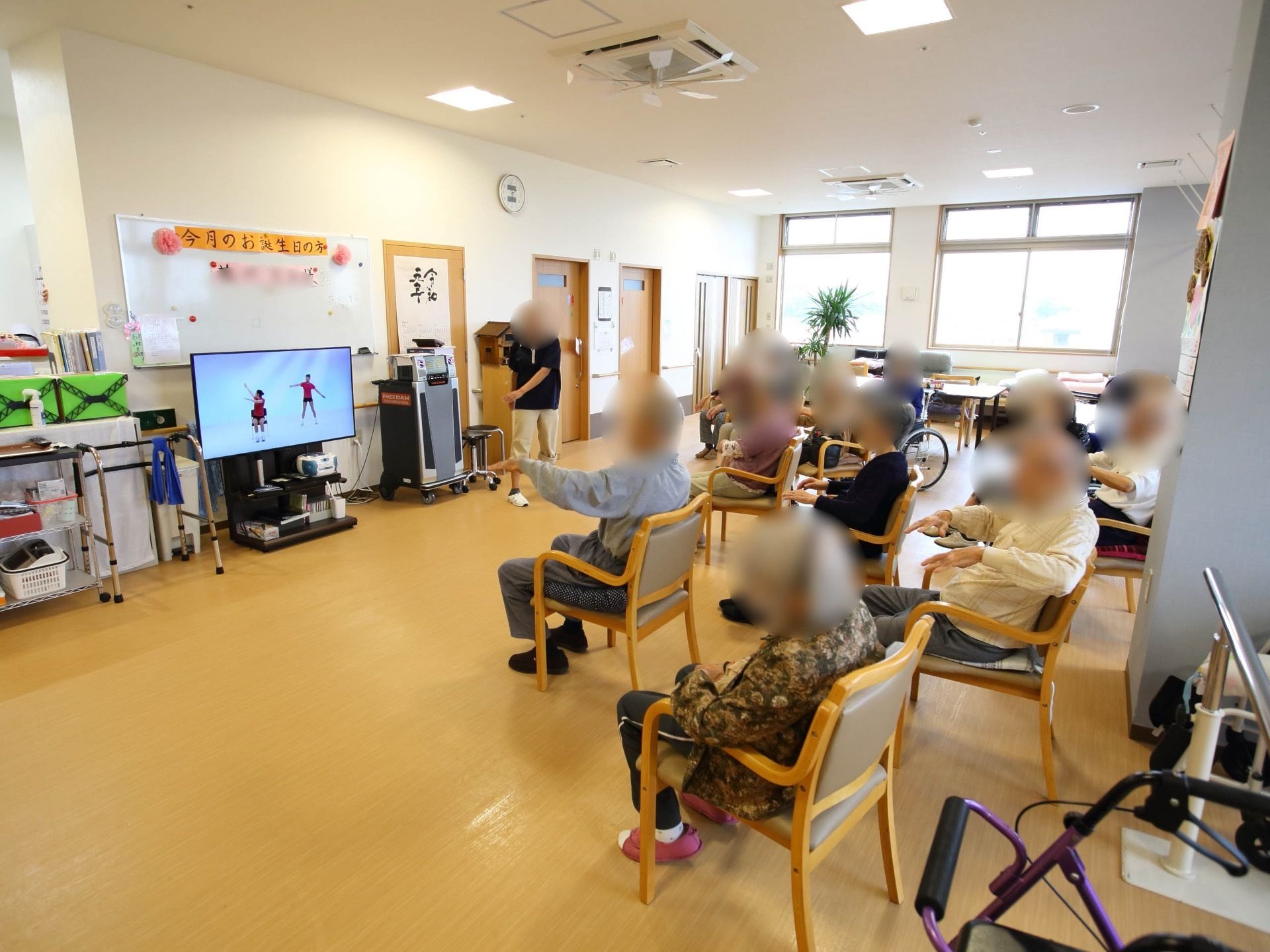 デイルームでは入居者様がテレビ画面を見ながら椅子に座って体操に取り組んでいる。部屋にはカラオケの機材もある。