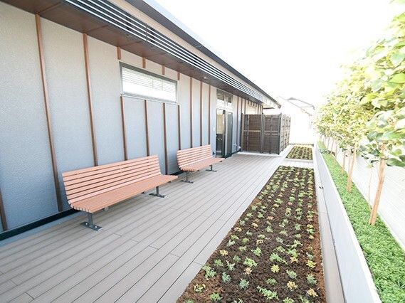 ウッドデッキが敷かれたテラスの前には草木が植えられた花壇があり、テラスには木製のベンチが二つ置かれている。奥には共有スペースにつながるドアがある、
