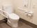 温水洗浄便座が取り付けられたトイレである。奥の壁にはＬ字型の手すりや非常ボタンが付けられている。