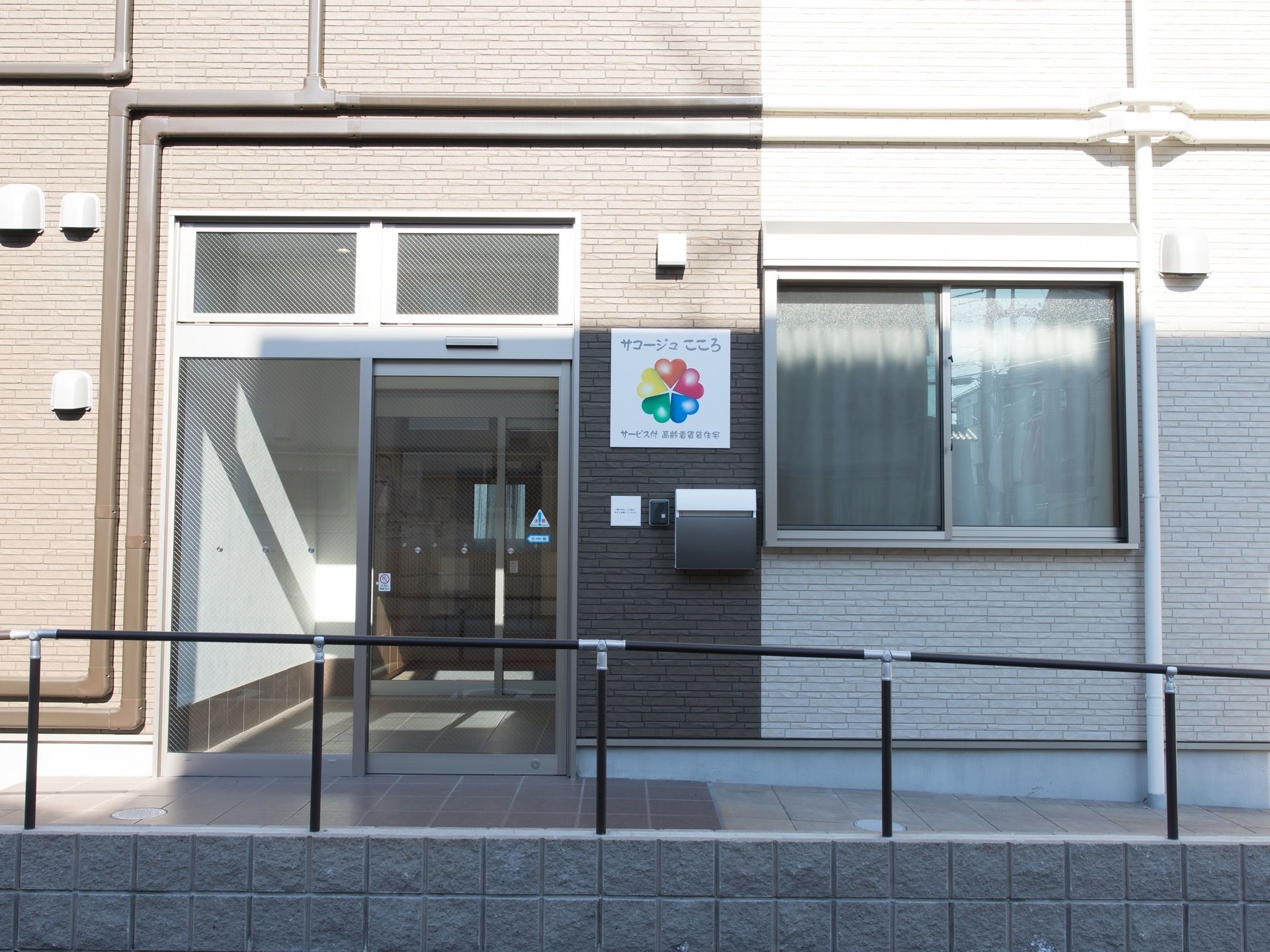 施設のマークが出入り口の目印である。手すりの設置された緩やかなスロープの先に、自動ドアが完備されている。