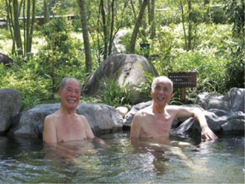 陽射しの中で露天風呂に浸かりこちらに向かって微笑む高齢の２人の男性