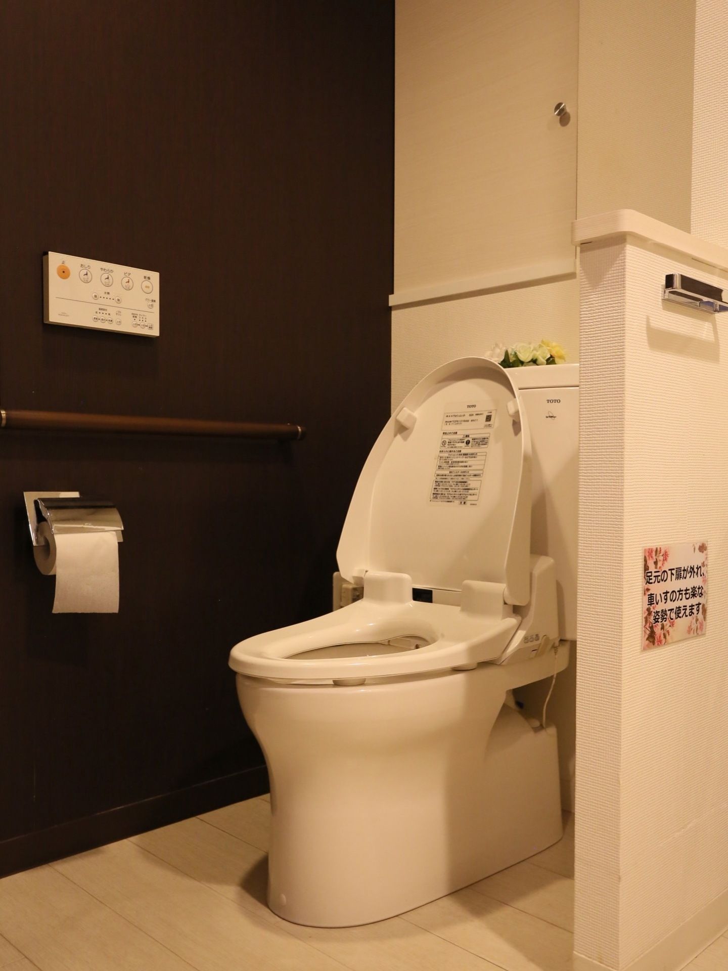 白いタンク式の洋式トイレの便座の蓋が開いている。茶色い壁にトイレのリモコンとペーパーホルダーと手すりが設置されている。