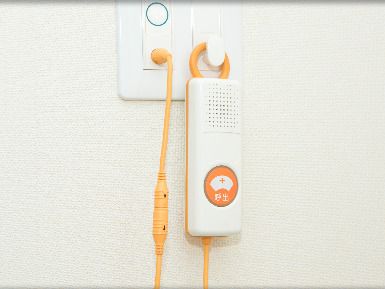 オレンジのコードが付いた緊急通報装置が付いた白い壁。ナース棒のイラストが緊急通報装置のボタンに描かれている。