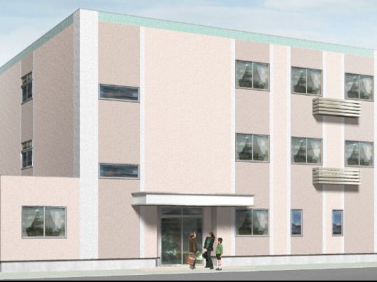 3階建ての建物。ベージュ系の外壁で玄関には屋根が付き、玄関前には3名の人がいて話をしている。