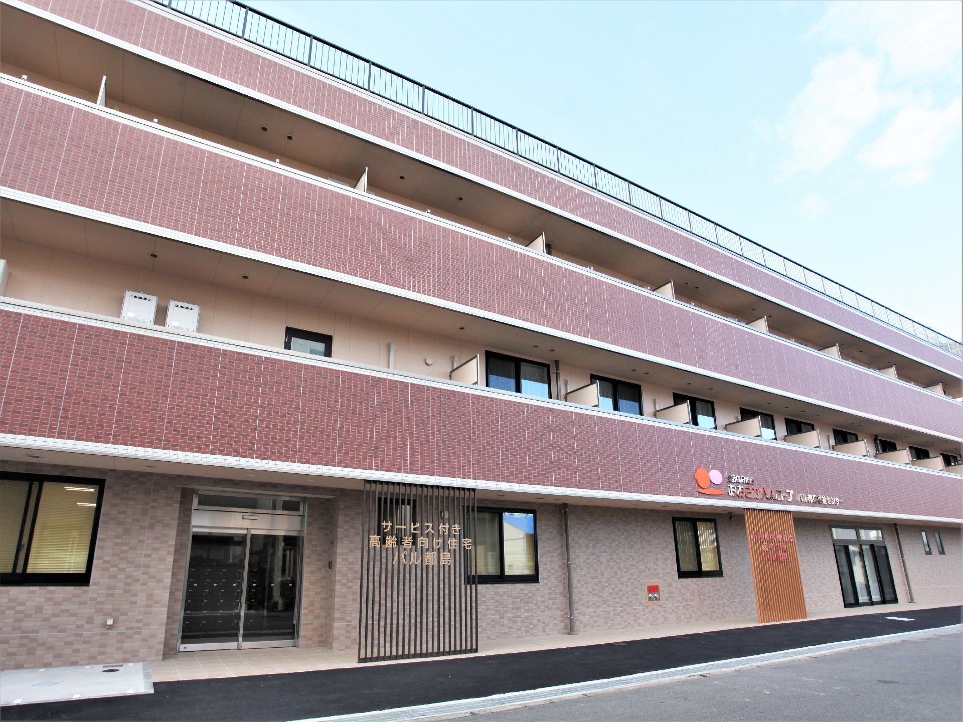 エンジ色の3階建ての建物。玄関付近には住宅の名前が書かれ、各フロアにベランダが付いている。