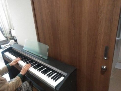 室内の茶色い扉の前で、座って電子ピアノを演奏する高齢者