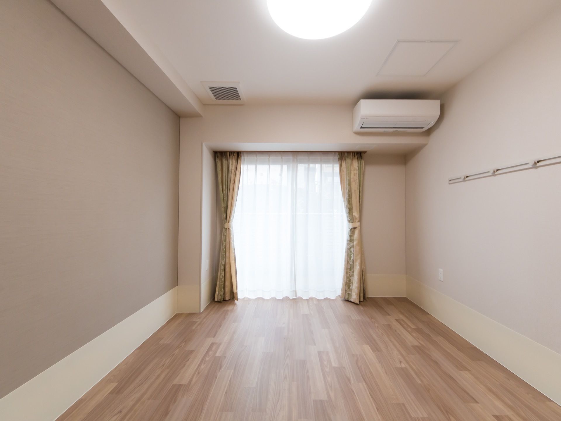 居室の奥には窓があり、明るい外光が差し込んでいる。その右上に、エアコンがある。家具などを置いていないスッキリした室内である。