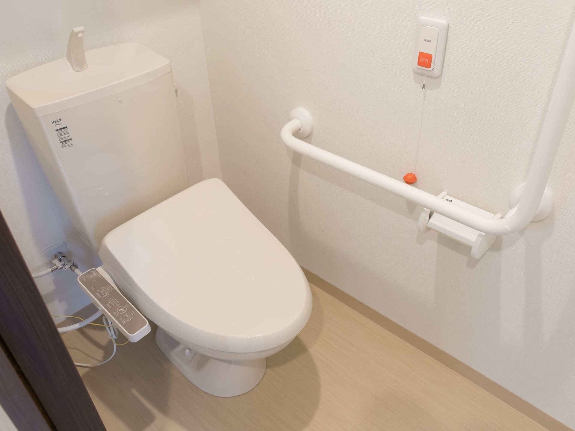 温水洗浄機能付きのトイレがある。壁には白くて大きなL字型の手すりが付いているおり、ナースコールが設置されている。