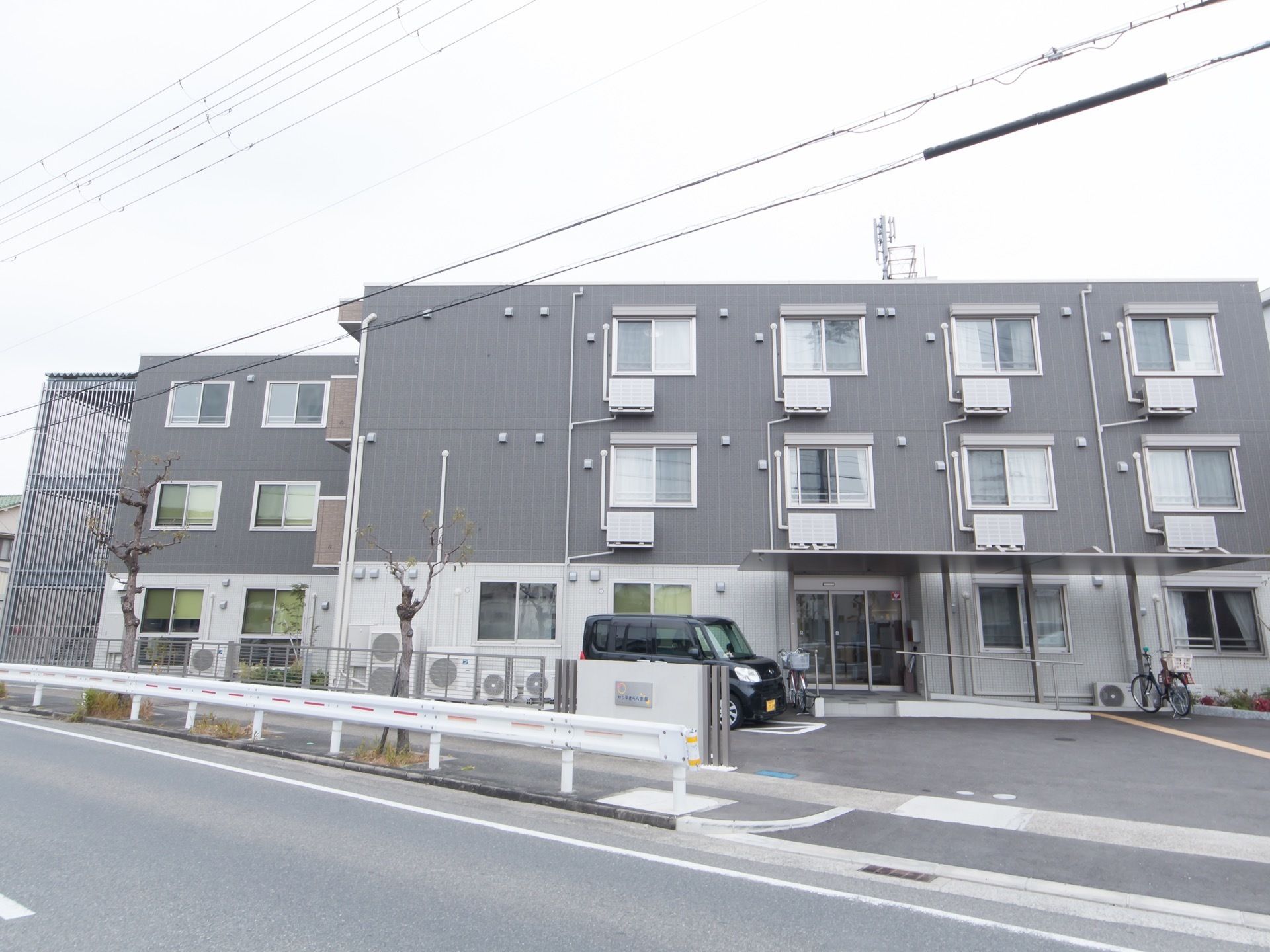 車道に面した敷地に3階建ての集合住宅が建っている。玄関部分には屋根があり、階段をはじめ手すり付きのスロープもある。