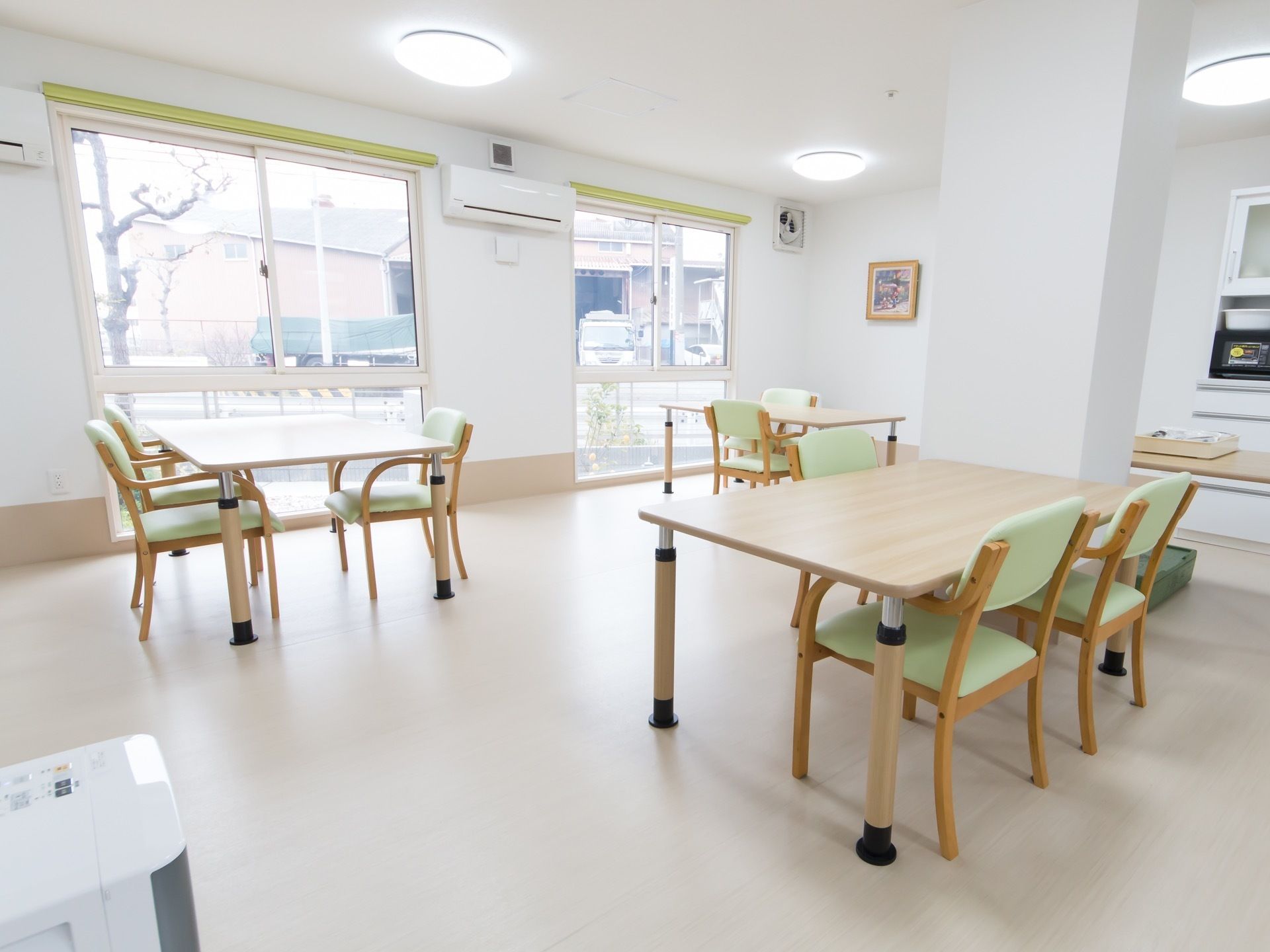 窓から外光の入る明るい食堂がある。テーブルセットが間隔を空けて並べられており、壁にはエアコンが設置されている。
