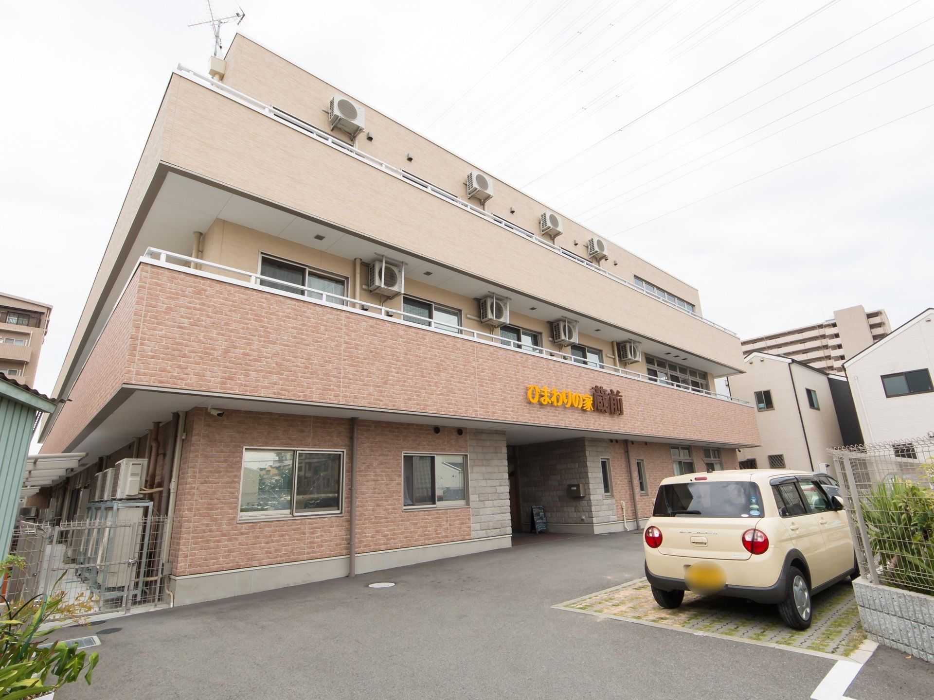 やさしい配色の3階建ての施設である。前面には、駐車スペースが設けられ、黄色い乗用車が1台停車している。