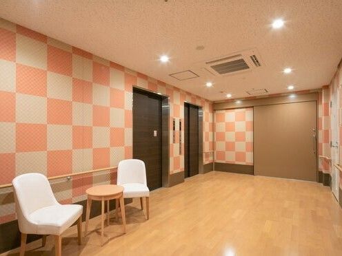 ピンクとベージュのチェック柄の壁紙が明るい雰囲気のエレベーターホール。白い椅子が小さな丸テーブルの両サイドに置かれている。